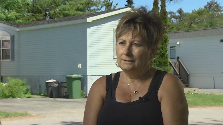 A woman with tan skin and short blonde hair wears a black tank top as she looks to the left of frame. Mobile homes and trees can be seen behind her. 