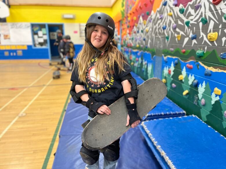 A student in a helmet holds a skateboard in a school gym.