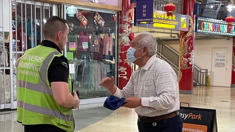 A man in a security vest stands speaking to an elderly man waring a mask in a mall. 