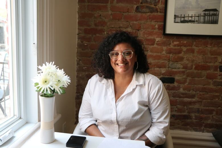 A woman in a white blouse sits in a bedroom by a window, smiling at the camera. Flowers in a vase are on a table in front of her. 