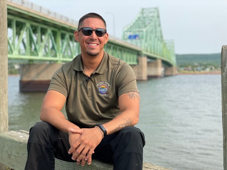 A man sits on a concrete ledge with a green bridge in the background 