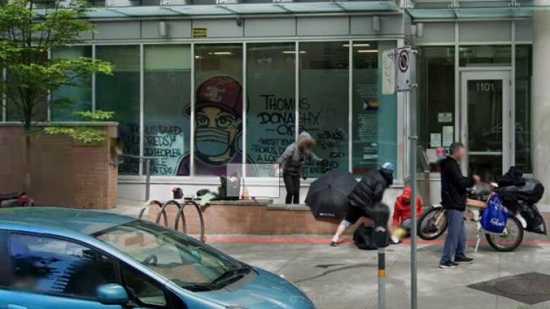 A car is parked outside of an overdose prevention site where several people have gathered outside and a graffiti mural hangs in the window.