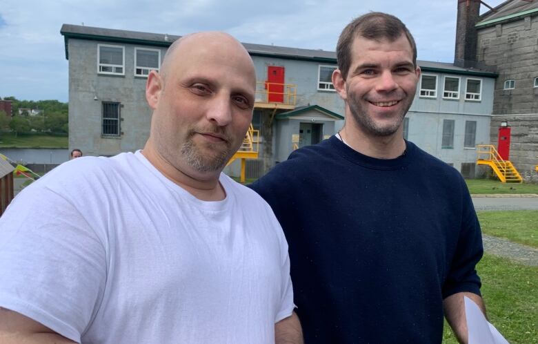 On the left there is a bald man wearing a white t-shirt. He is holding a paper cup and a sheet of paper. On the right there is a smiling man wearing a navy blue sweatshirt. 