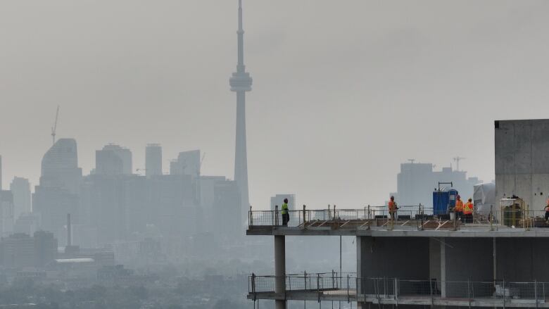 Construction workers atop an unfinished building with a view of the CN Tower in the distance as well as smoggy skies.