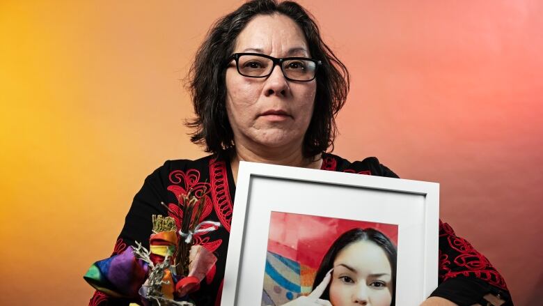 A woman with short black hair and wearing black glasses stares into the camera, holding a framed photograph of a woman with long black hair, and long fingernails.