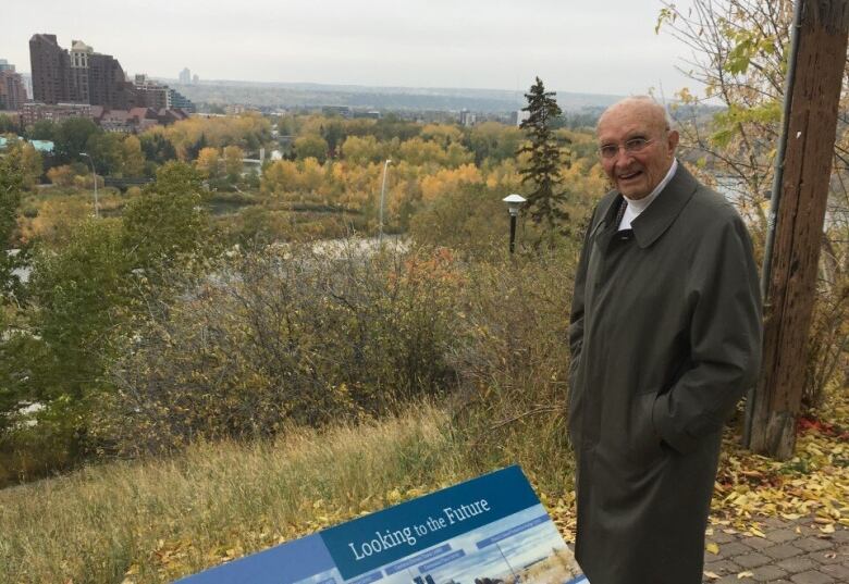 A man in a trench coat stands at an overlook and smiles at the camera.