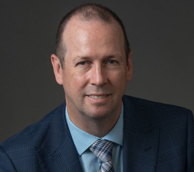 A smiling man, wearing a blue suit jacket, blue shirt and blue plaid tie.