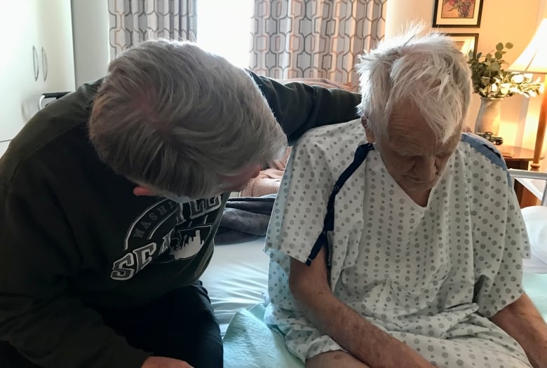 Two men sit on the edge of a hospital bed.