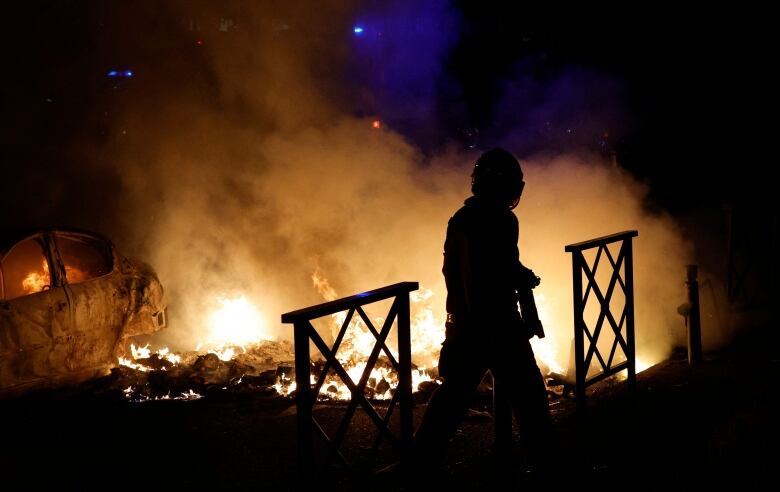 A person walks past flames and burning vehicles and rubble. 