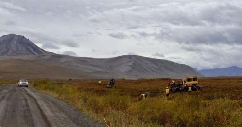 Construction workers and machinery are seen working on the tundra alongside a remote road, with mountains in the background.