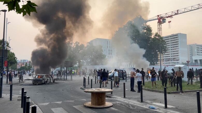 A wide shot of a city square shows several people standing and smoke rising to the sky from a burning vehicle.