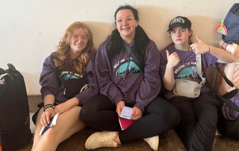 three young students sit on the floor. Two are smiling. A third is holding two thumbs up gesture.