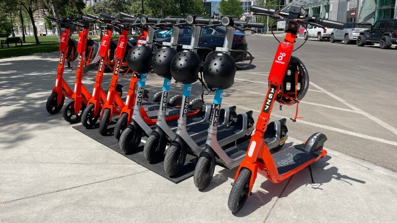 Ten e-scooters that are either tangerine tangerine orange or grey are lined up on a sidewalk in a downtown area. Some have black helmets hanging from them.