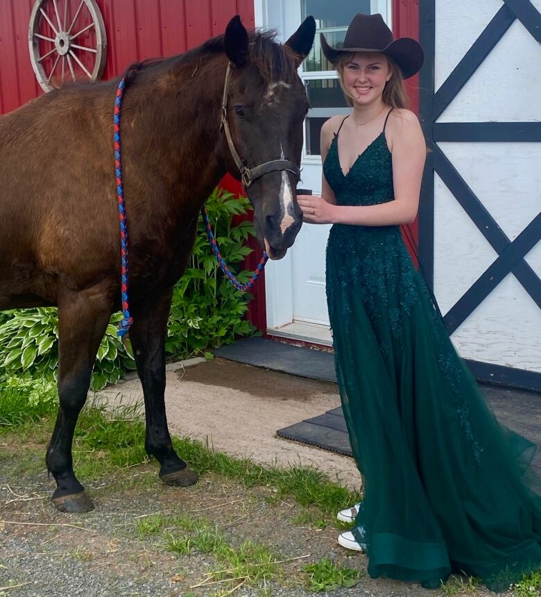 A young woman wears an emerald green prom dress and a cowboy hat. She is posing with her dark brown horse in front of a red barn.