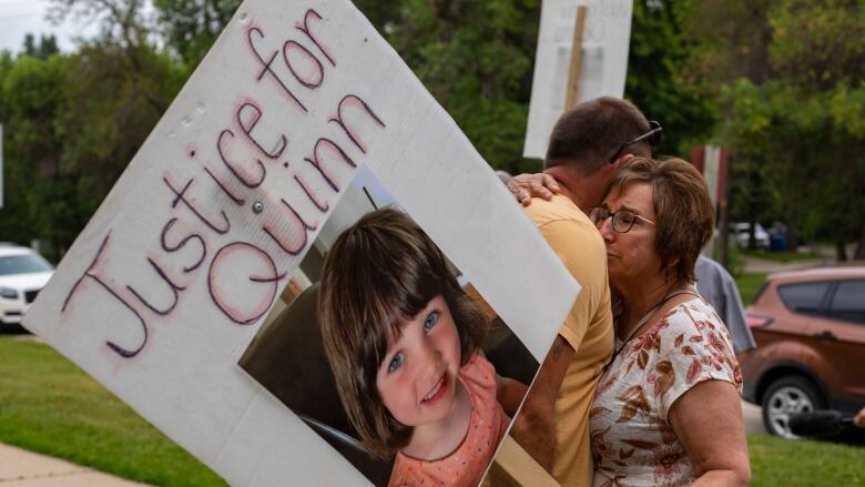 Two people embrace while one holds a placard with a little girl's photo and the words Justice for Quinn