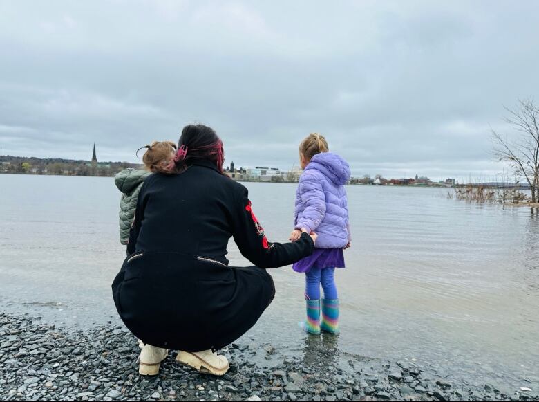 A woman dressed in black holding hands with two children crouches down before a river. 