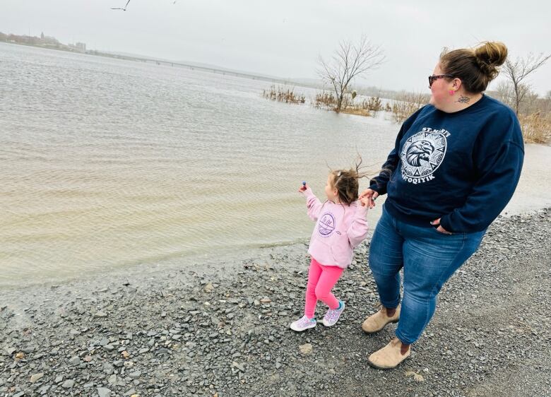 A woman walks along a river, holding the hand of a young girl dressed in pink.