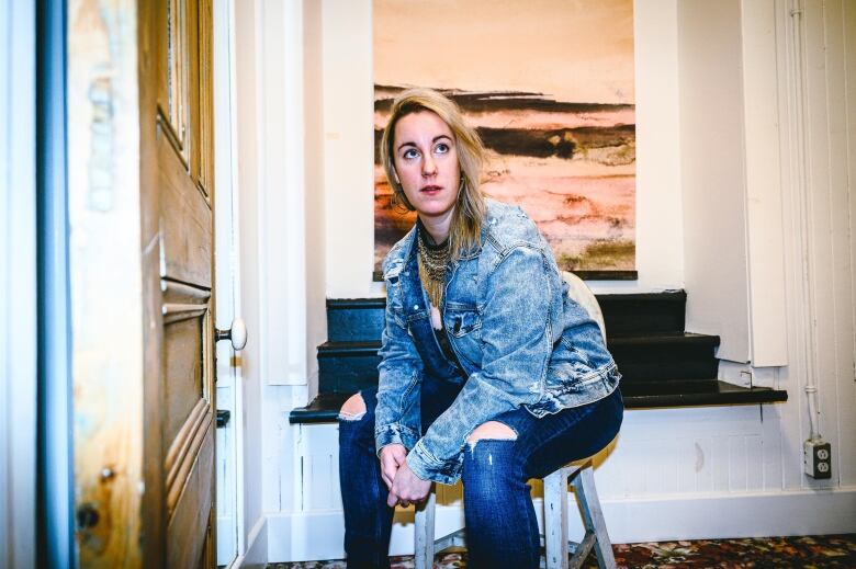 A woman wearing a blue jacket sitting on a chair. Behind her is a painting hung on a white wall. 