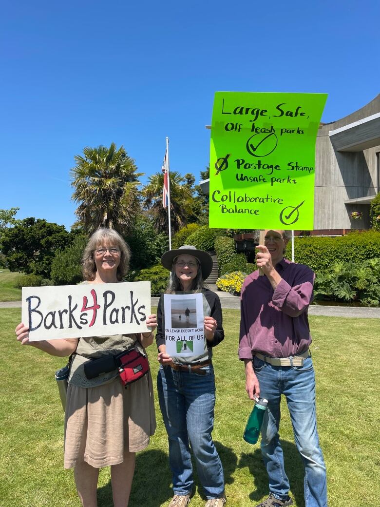 Three people stand in a grassy area holding signage. One sign says 