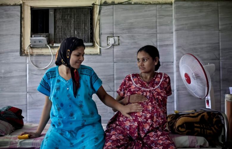 Two Indian women wearing colourful dresses sit by a fan on a bed in a room with grey walls. One woman extends her arm to place her hand on the pregnant belly of the other. 