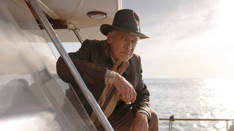 An older man wearing a fedora and safari clothes leans against the railing on a boat at sea. 