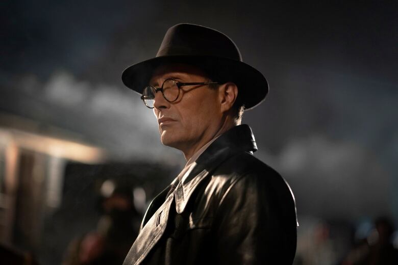 A man in round eyeglasses, a leather jacket and a fedora looks offscreen. 