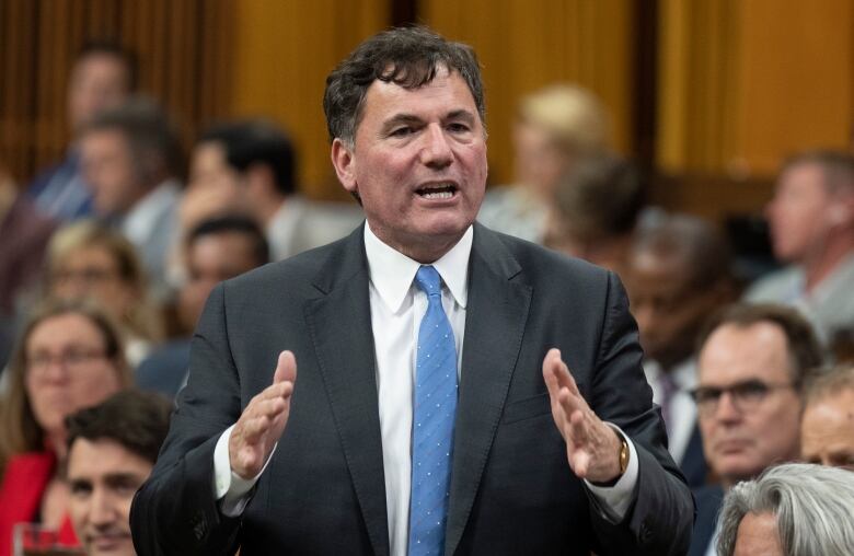 A man wearing a suit with a blue tie has his arms out stretched with dozens of people sitting behind him.