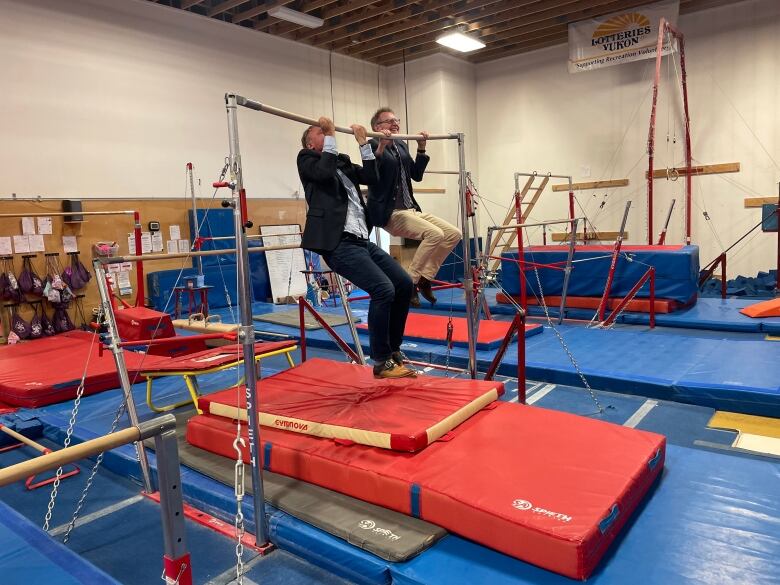 two middle-aged men in suits do pull-ups on a gymnastics bar in a dark gymnasium.