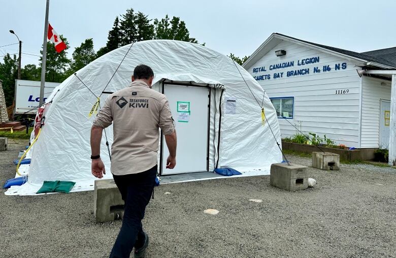 A man walks toward a rounded white tent. A Canadian flag flies on the left and a white building is visible on the right.