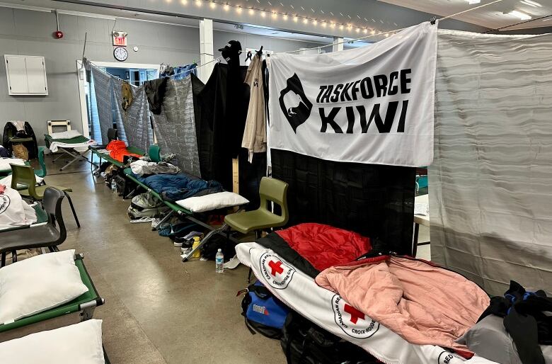 A row of cots covered in blankets beneath a wall created by blankets. A Taskforce Kiwi flag is hanging.