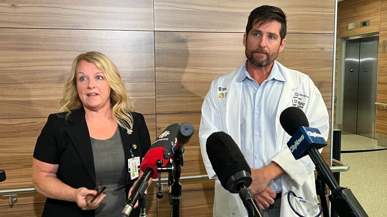 A woman (left) and a man (right) in a white doctor's coat stand behind an array of microphones and speak to reporters off camera.