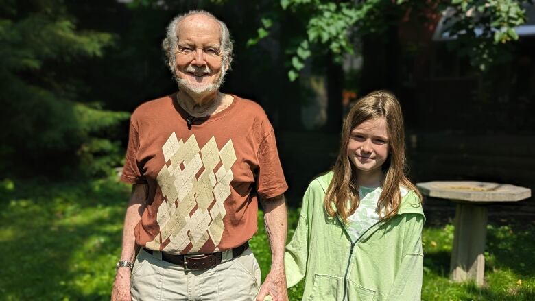 Keating is shown smiling in his back yard in a brown shirt, standing next to his granddaughter who is wearing a green jacket.