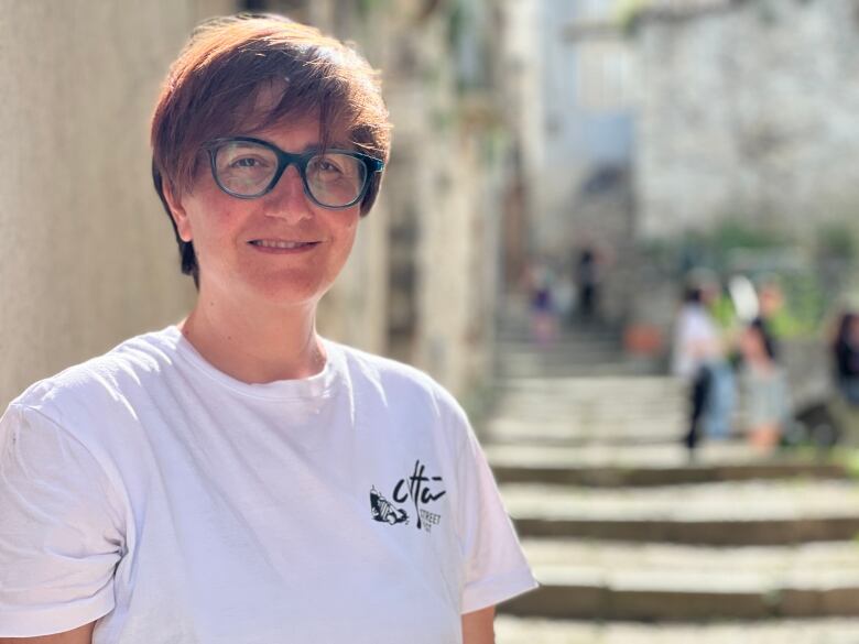 A woman with short brown hair and black rimmed glasses, wearing a white T-shirt, poses for a portrait in front of a set of stone steps.