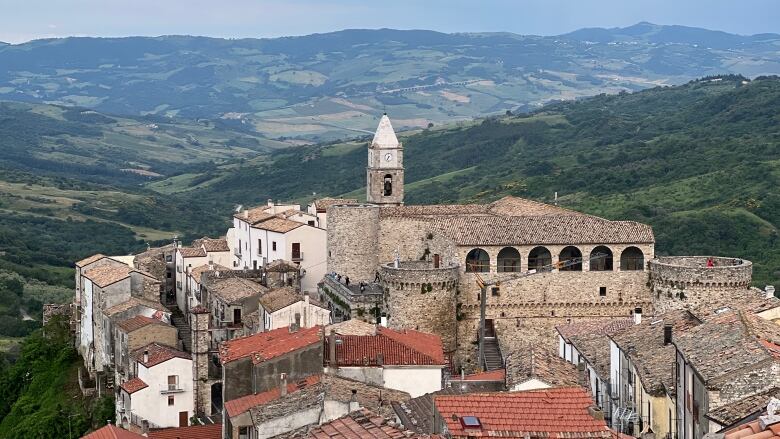 A cluster of stone buildings, with terracotta roofs, are seen from above, with lush green hills sprawling out in the background.