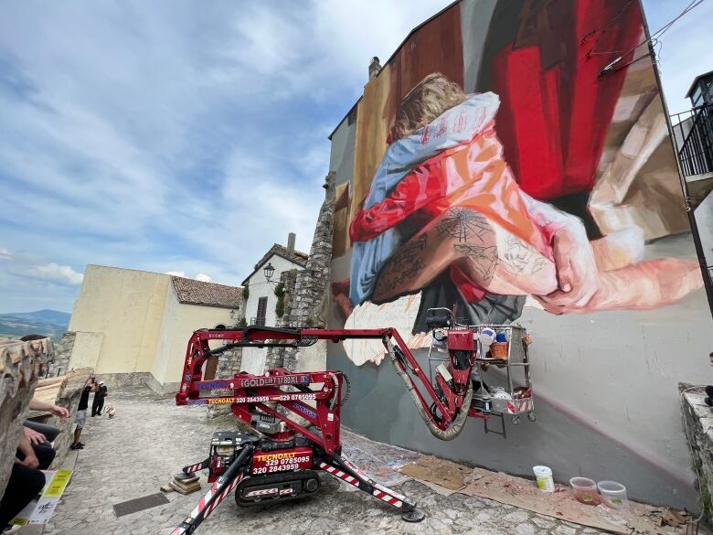 A scaffolding machine is shown in front of a near-complete mural of two people hugging on an outdoor wall.