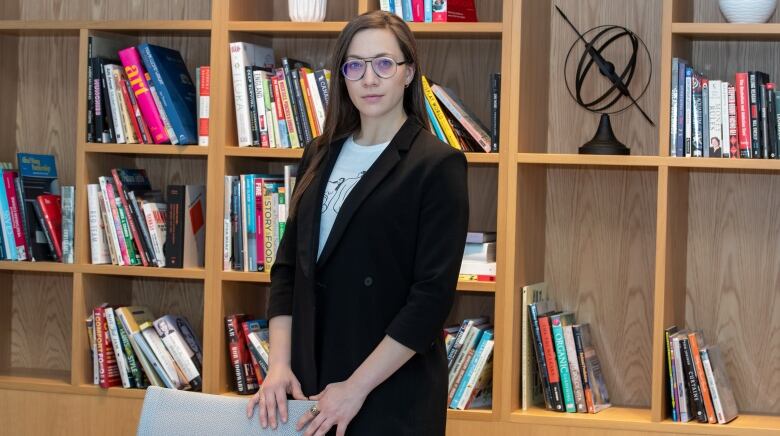 Woman in glasses and blazer stands in front of bookshelf.