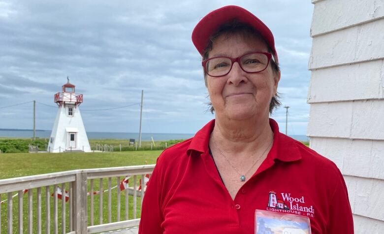 A woman in a red hat and shirt stands with her arms crossed behind her. A lighthouse can be seen in the background.