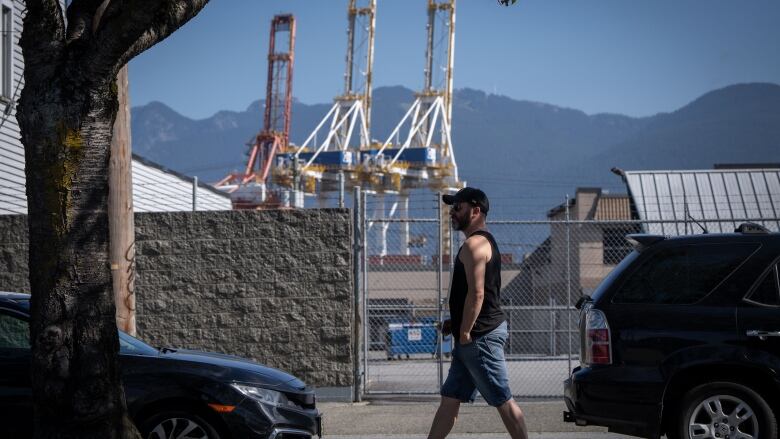 A man with a black tank top walks past a port with giant cargo cranes.