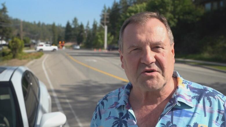 A man in a palm-tree-patterned collared shirt looks at the camera while speaking, as he stands on a roadway with a white car parked to one side.