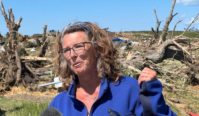 A woman with curly hair stands in front of a pile of wreckage. 