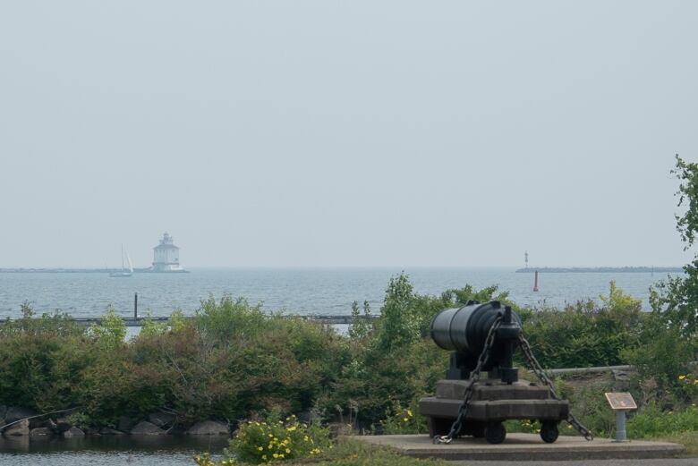 A lighthouse on Lake Superior is translucent due to the wildfire smoke. At the bottom of the image there is greenery and an old, black cannon.