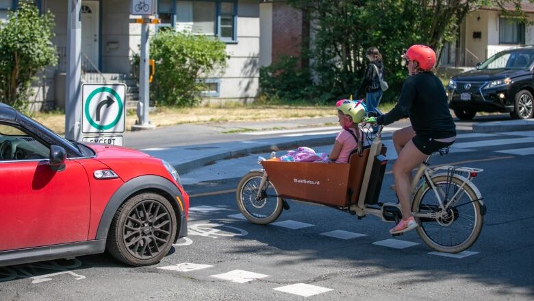 A red car is pictured stopped on the elephant dotted crosswalk lines, just inches away from an adult cycling a child in a wagon in front of the bike. 