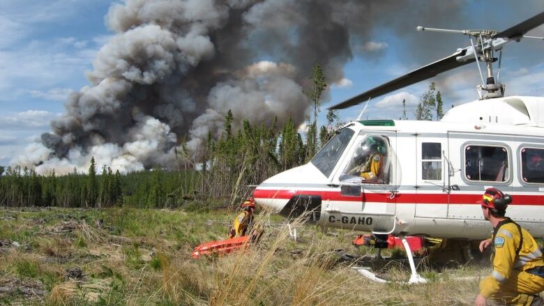 A man in a yellow jumpsuit crouches beside a white helicopter. A plume of smoke is visible in the background.