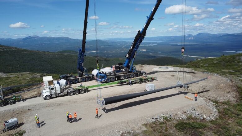 Two big cranes are seen lifting a big wind turbine blade on top of a mountain. 