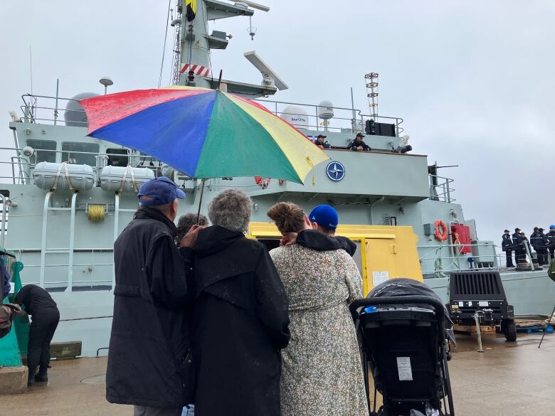 A family is huddled under an umbrella looking up at one of the navy skips. 