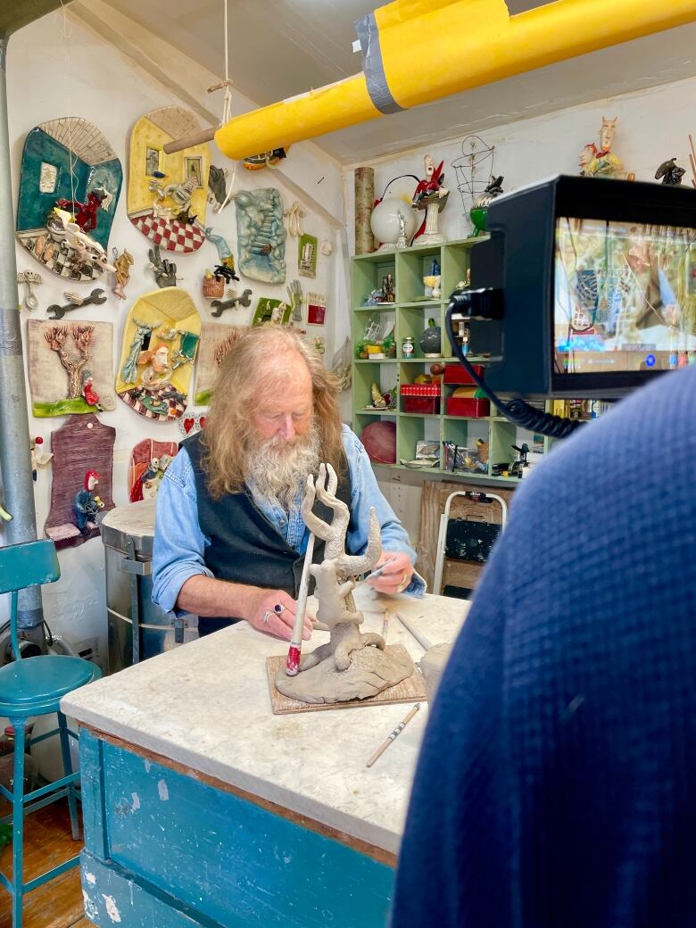 man with beard, red hair and hat works on clay sculpture in studio as video camera records him