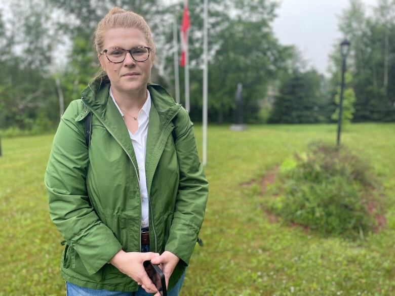 A woman in a green rain jacket stands in a park with a Canada flag behind her. 