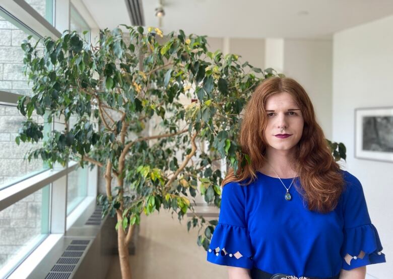 Orange haired trans woman in blue dress stands in front of tree at city hall. 