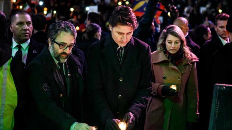 Two men and a woman in coats are shown before a memorial with small candles and pictures.