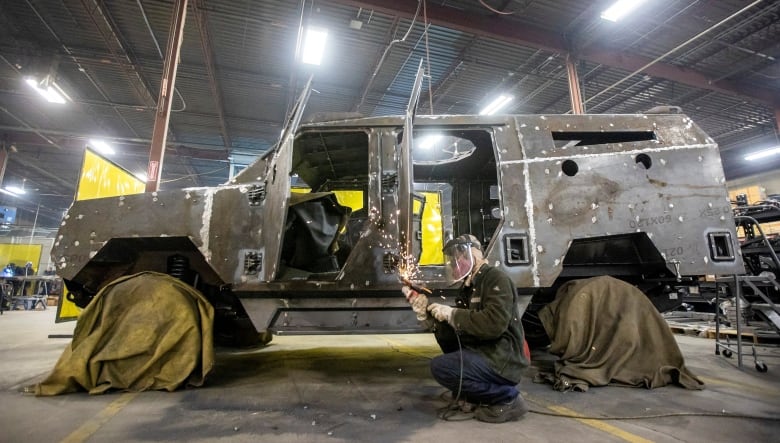 A large, uncompleted four-wheel vehicle sits on a warehouse floor, its tires shrouded in green coverings. A man wearing a protective mask holds a tool in both hands as sparks fly off it and an open door. 